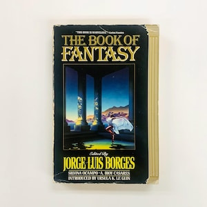 Może przedstawiać: Książka w twardej oprawie zatytułowana "THE BOOK OF FANTASY" z czarną okładką i złotymi literami. Okładka przedstawia surrealistyczną scenę z postacią w białej tkaninie. Zredagowane przez Jorge Luis Borges.