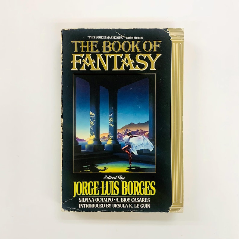Może przedstawiać: Książka w twardej oprawie zatytułowana "THE BOOK OF FANTASY" z czarną okładką i złotymi literami. Okładka przedstawia surrealistyczną scenę z postacią w białej tkaninie. Zredagowane przez Jorge Luis Borges.
