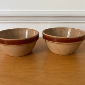 Puede incluir: Dos cuencos pequeños de cerámica marrón con un borde marrón. Los cuencos están sobre una superficie de madera.