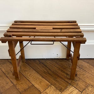 Puede incluir: Un taburete pequeño de madera con una superficie superior de listones construida con varias tablas horizontales. La estructura se apoya en cuatro patas e incluye una barra de soporte de metal oscuro debajo del asiento.