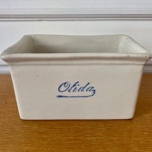 Peut inclure: Un récipient rectangulaire en céramique blanc cassé avec un bord légèrement évasé. Le mot "Olida" est écrit en script bleu sur le devant. Le récipient est posé sur une surface en bois.