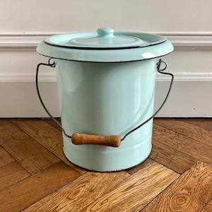 May include: A vintage aqua enamelware bucket with a lid. The bucket has a black rim and handle attachments, with a wooden handle. The lid has a small knob. The bucket is sitting on a wooden floor.