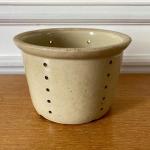Peut inclure: Un bol à baies en céramique beige avec une finition lisse et émaillée et un bord large et arrondi. Le bol présente des rangées verticales de petits trous de drainage régulièrement espacés sur les côtés pour la ventilation et l'égouttage.