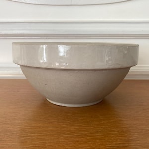 Peut inclure: Un bol en céramique beige clair avec un large bord. Le bol a une forme arrondie et repose sur une surface en bois. L'arrière-plan présente un mur blanc avec des moulures.