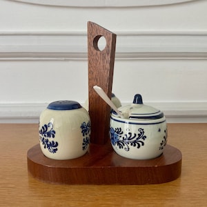 May include: A wooden salt and pepper shaker holder with a blue and white floral design. The holder has a small spoon and a lidded jar.