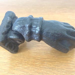 Peut inclure: Un heurtoir de porte en fonte noire en forme de poing fermé. Le heurtoir est monté sur une plaque rectangulaire avec une finition rouillée. Le poing est détaillé avec des jointures et des rides.