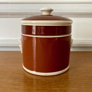 Peut inclure: Un pot en céramique marron avec un couvercle, avec un bord blanc et une poignée décorative sur le dessus. Le pot a une finition brillante et deux petites poignées décoratives sur les côtés. Le pot est posé sur une surface en bois.