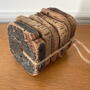 May include: A set of six cork coasters bound with twine. The coasters are made from natural cork, displaying a dark, textured pattern. The edges are curved, and the twine is light brown. The coasters are on a wooden surface.
