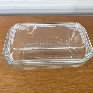 以下が含まれることがあります： 蓋に牛のデザインが施された透明なガラス製のバター入れ。長方形で、滑らかで透明な仕上がりです。