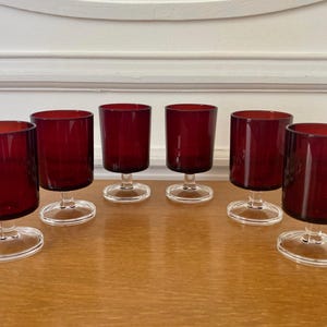 Peut inclure: Six verres à vin rouge rubis vintage avec des bases ovales transparentes. Les verres ont une forme cylindrique et sont disposés sur une surface en bois. L'arrière-plan est un mur blanc avec une garniture décorative.