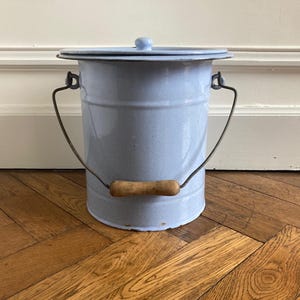 May include: A light blue enamel bucket with a matching lid and a metal swing handle featuring a cylindrical wooden grip. The bucket has a cylindrical body with a slightly flared rim and a small knob on the center of the lid for easy removal.