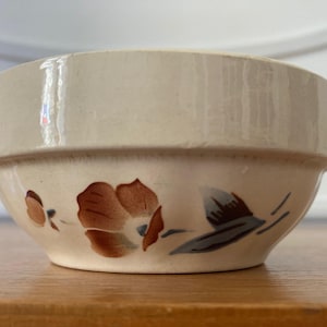 Puede incluir: Cuenco de cerámica color crema con un diseño floral. El cuenco presenta un borde ancho y una forma ligeramente acampanada. El diseño floral incluye flores marrones y azul-gris, añadiendo un toque de encanto rústico.