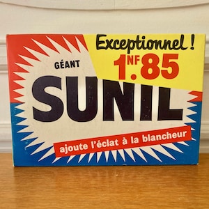 May include: Vintage SUNIL laundry detergent box. The box features a bold design with the word "SUNIL" in large, dark letters. The box has a red, yellow, and blue color scheme with text in French. The box is on a wooden surface.