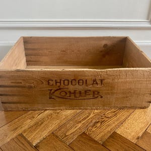 May include: A rectangular wooden crate with the words "CHOCOLAT KOHLER" printed in dark red. The crate is made of light brown wood and sits on a wooden floor.