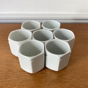 以下が含まれることがあります： 7つの白い六角形のセラミックカップのクラスター。カップは木製の表面に円形に配置されています。カップは滑らかで光沢のある仕上がりで、シンプルでモダンなデザインです。背景は白い壁です。