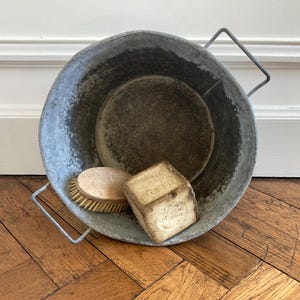 May include: A circular, aged metal basin with wire handles, holding a wooden scrubbing brush, a round wooden item, and a bar of soap. The basin has a grey, worn look, indicating vintage use. The contents suggest a cleaning or laundry theme.