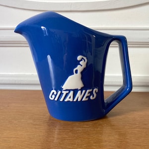 May include: A cobalt blue ceramic pitcher with a unique, angled design. The pitcher features a white logo and the word "GITANES" in white lettering. It has a rectangular handle and a spout for pouring.