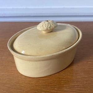 Peut inclure: Un plat en céramique de couleur crème, de forme ovale, avec un couvercle assorti. Le couvercle est orné d'un bouton décoratif et texturé. Le plat est posé sur une surface en bois, suggérant un cadre de cuisine ou de salle à manger.