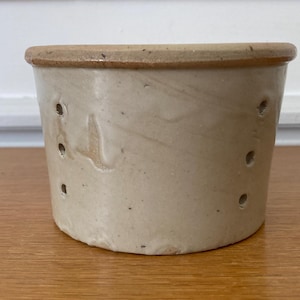 Peut inclure: Un récipient cylindrique en céramique blanc cassé avec un bord marron clair. Le récipient comporte plusieurs petits trous espacés régulièrement sur le côté. La surface présente de subtiles variations de couleur et de texture.