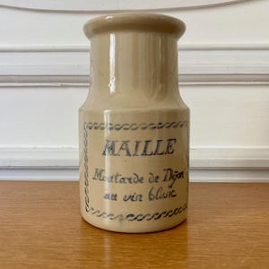 May include: A beige ceramic jar with a cylindrical shape. The jar features blue text that reads "MAILLE Moutarde de Dijon au vin blanc." The jar is sitting on a wooden surface.