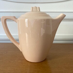 May include: A vintage, peach-colored ceramic teapot with a lid and a curved handle. The teapot has a spout and sits on a wooden surface. The lid has a decorative floral design.