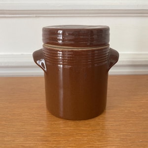Op de afbeelding: Een bruine keramische pot met een deksel en twee kleine handvatten. De pot heeft een cilindrische vorm en is ongeveer 13 cm hoog. Het deksel sluit goed aan en de pot heeft een glanzende afwerking.