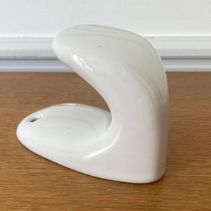 Op de afbeelding: Een glanzende witte keramische wandhaak met een glad, gebogen, J-vormig ontwerp. De basis is voorzien van twee ronde montagegaten voor installatie en de algehele vorm is afgerond en solide, wat zorgt voor een strakke, reflecterende afwerking.