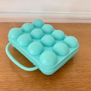 Puede incluir: Un recipiente rectangular de plástico color turquesa claro para almacenar huevos, con una rejilla moldeada de doce compartimentos redondeados para huevos individuales. Incluye un asa integrada resistente en un lado.