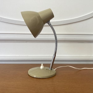 May include: A beige desk lamp featuring a conical shade, a flexible chrome-finished gooseneck arm, and a circular base. The lamp includes a white push-button switch located on the base and a white electrical cord extending from the rear.