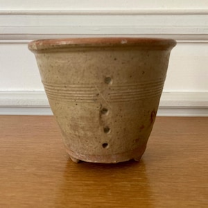 May include: A small, antique-style, tan ceramic pot with a slightly flared shape. It has horizontal etched lines and three small holes. The rim is a darker brown colour.