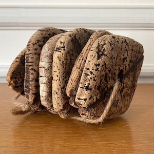 May include: A rustic, handmade cork object with a natural, textured appearance. The cork is a light brown color with dark brown and black markings. A natural twine is wrapped around the object. The object is on a wooden surface.