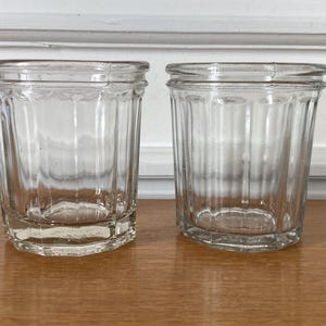 May include: Two clear glass jars with vertical ribbed detailing along the sides and a thick, reinforced rim at the top. Each jar features a wide opening and a sturdy, flat base, showcasing a transparent design that highlights the glass texture.