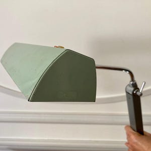 May include: A desk lamp featuring a pale green, angular metal shade with a small red and yellow switch on top. The shade is attached to a chrome-plated arm that connects to a dark grey, rectangular vertical stand with a silver adjustment lever.