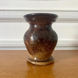 Op de afbeelding: Een kleine, handgemaakte keramische vaas met een bolvormig lichaam en een smalle hals. De vaas heeft een donkerbruin glazuur met lichtere bruine en tan accenten, waardoor hij een rustieke uitstraling heeft. De vaas staat op een houten oppervlak.