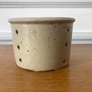 Pode incluir: Um molde de queijo cilíndrico de cerâmica com um acabamento bege salpicado. O recipiente apresenta uma borda plana e vários pequenos orifícios de drenagem circulares dispostos em filas verticais nas paredes externas, exibindo uma aparência terrosa.