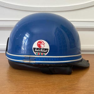 Può includere: Un casco da moto a mezza calotta blu lucido con un adesivo circolare bianco raffigurante un'icona di casco rosso e la scritta Jumbo Helmet sopra un piccolo motivo a strisce verdi, bianche e rosse. Un bordo testurizzato bianco e blu circonda la base.