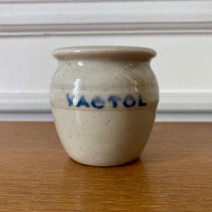 Puede incluir: Un pequeño tarro de cerámica blanquecina con forma redondeada y un borde ligeramente ensanchado. La palabra "VACTOL" está impresa en azul en la parte delantera. El tarro está sobre una superficie de madera.