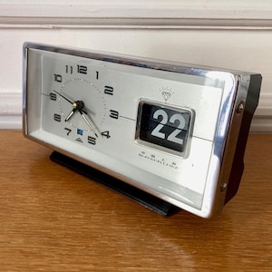 Könnte beinhalten: Eine silberne und schwarze Vintage-Klappuhr mit rechteckigem Zifferblatt. Die Uhr zeigt die Zeit mit schwarzen Zeigern und Ziffern auf silbernem Hintergrund an. Das Datum wird als "22" in einem schwarzen Feld angezeigt. Die Uhr ist mit "SHANGHAI CHINA" beschriftet.