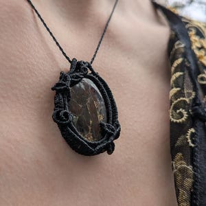 May include: A black macrame necklace featuring an oval-shaped, dark brown stone with gold flecks. The stone is encased in a woven black frame and hangs from a delicate black cord. The necklace is worn against a person's skin.