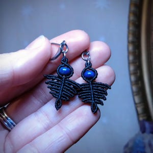 Peut inclure: Une paire de boucles d'oreilles en macramé noir au design unique. Chaque boucle d'oreille présente une perle ovale bleu foncé en haut, avec un motif en forme d'éventail en dessous. Les boucles d'oreilles ont des crochets d'oreilles argentés.