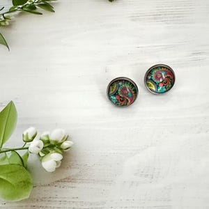 Peut inclure: Une paire de boucles d'oreilles à clous rondes présentant un motif cachemire multicolore et vibrant sous un dôme en verre transparent. Les boucles d'oreilles sont montées sur une monture en métal sombre, avec des tourbillons de rose, vert, bleu et jaune.