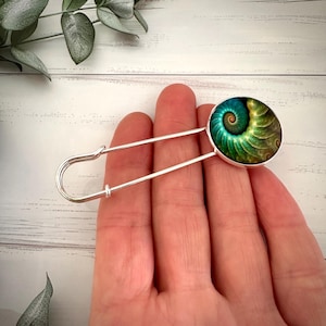 May include: A silver safety pin with a round green and gold patterned glass cabochon. The cabochon features a spiral design.