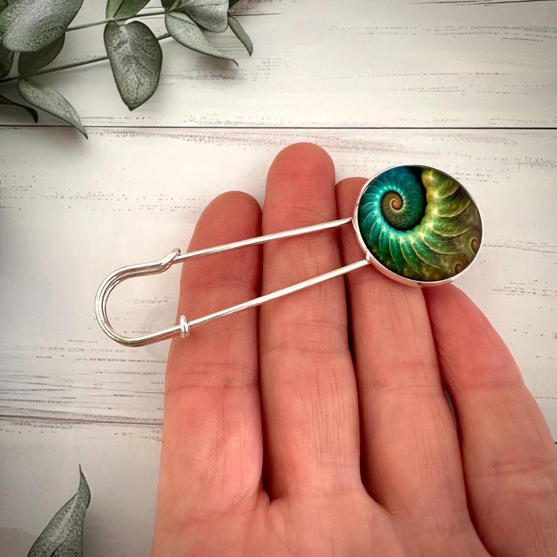 May include: A silver safety pin with a round green and gold patterned glass cabochon. The cabochon features a spiral design.