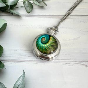 May include: A silver pocket watch with a chain, featuring a vibrant spiral design in shades of green, blue, and yellow. The watch is set against a white wooden background with green foliage.