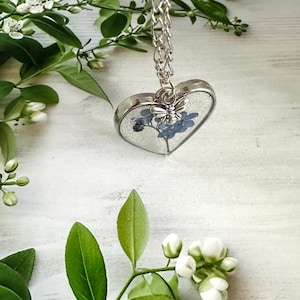May include: A silver heart-shaped pendant necklace with a delicate silver chain. The pendant contains a pressed forget-me-not flower and a small silver butterfly charm.
