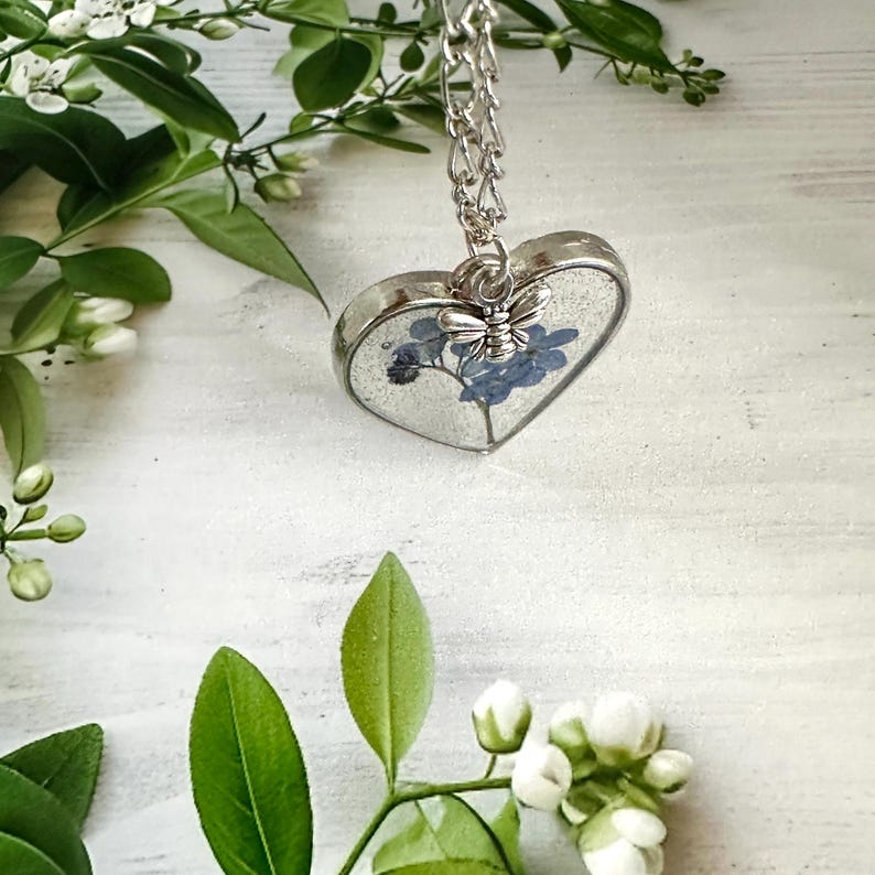 May include: A silver heart-shaped pendant necklace with a delicate silver chain. The pendant contains a pressed forget-me-not flower and a small silver butterfly charm.