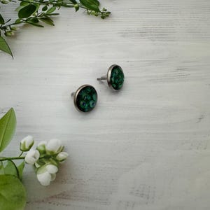 May include: A pair of stud earrings with a silver setting. The earrings have a round, dark green design with blue accents, reminiscent of peacock feathers. They are displayed on a white wooden surface with floral accents.