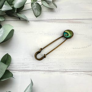 May include: A gold safety pin with a green and blue circular glass charm featuring a spiral design.
