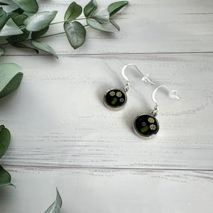 May include: A pair of silver-toned dangle earrings. Each earring features a small, round, black resin pendant with tiny white flowers and green accents. The earrings are displayed on a white wooden surface with green foliage in the background.