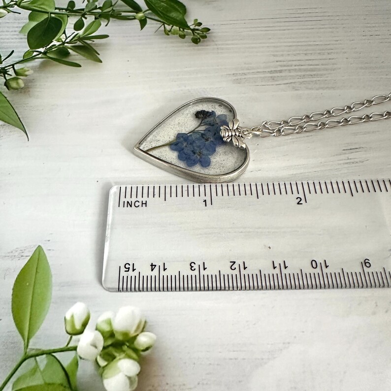 May include: A silver heart-shaped pendant necklace with a clear resin insert containing pressed blue forget-me-not flowers. The pendant is approximately 1 inch in diameter and hangs from a silver chain.